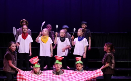 children with heads poking out of tablecloth and porridge bowls on their heads plus children in white t shirts and holding wooden spoons 